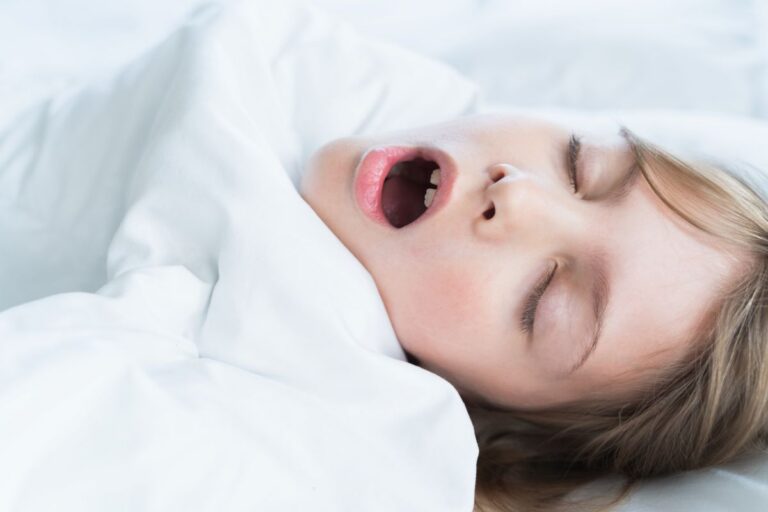 Person yawning while lying in bed under a white duvet, eyes closed and mouth open