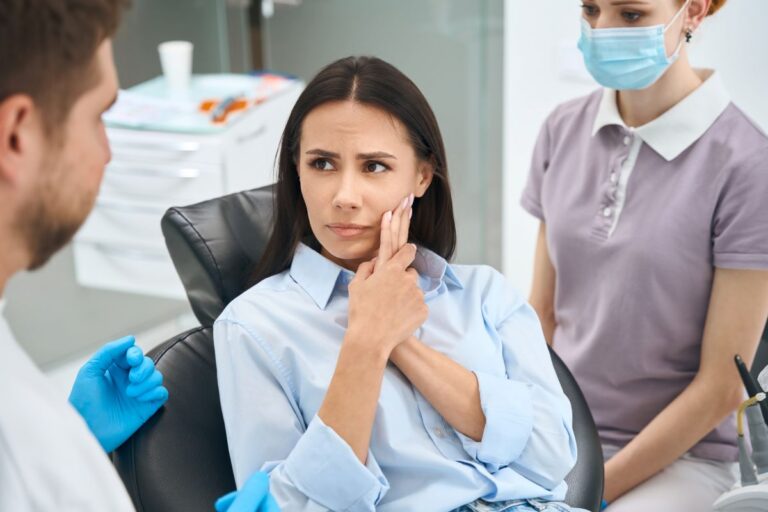 Woman in a light blue shirt sits in a dental chair, pressing her jaw in apparent pain while a clinician in blue gloves talks nearby.